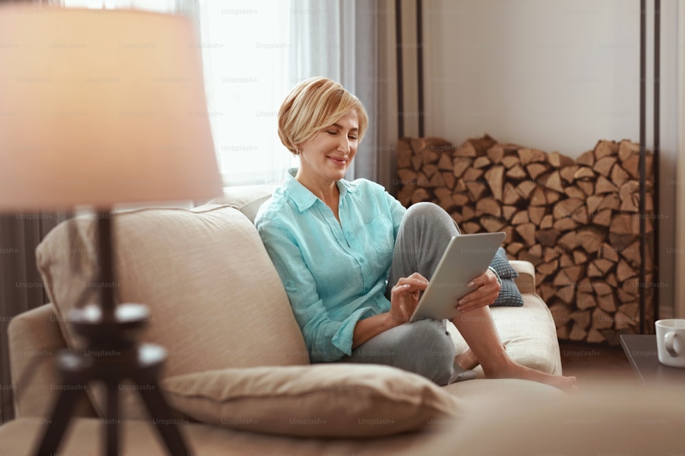 Female With Tablet. Mature Woman On Sofa In Beige Plaid Uses Digital Device Touchscreen, Comfortable Internet Search. High Resolution.