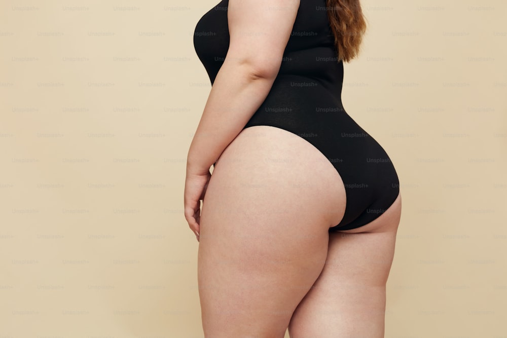 플러스 사이즈 모델. 여자는 엉덩이를 닫습니다. 검은 바디수트를 입은 뚱뚱한 몸통. 베이지색 배경에 포즈를 취하는 완전한 모습의 여성. 바디 포지티브 개념.