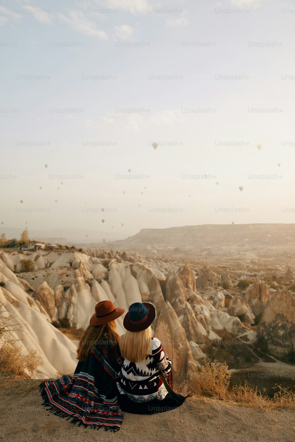 Travel. Women Travelers Looking At Flying Hot Air Balloons In Sky, Female In Hats Sitting On Hill Enjoying Sunset. High Resolution