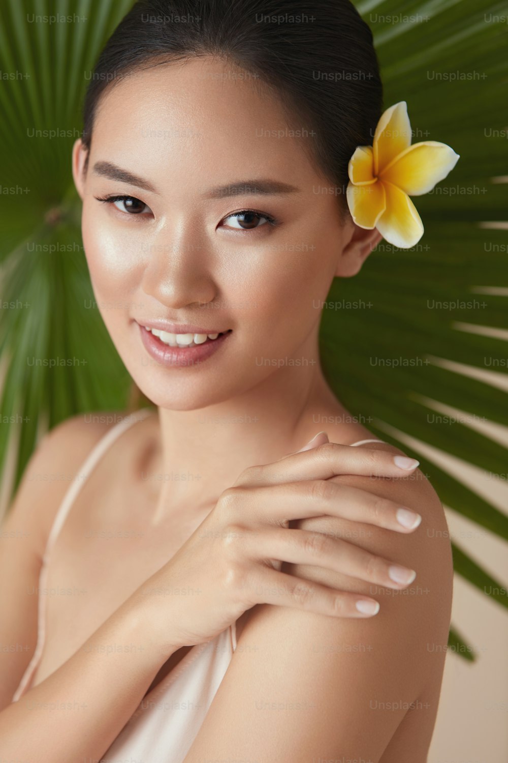 Beauté. Modèle près de la palme avec fleur dans les cheveux. Tendre femme asiatique touchant l’épaule portrait sur fond de plante tropicale. Belle fille profitant d’une peau hydratée après avoir utilisé des cosmétiques biologiques naturels.