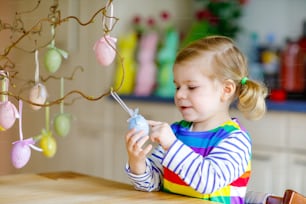 Cute little toddler girl decorating tree bough with colored pastel plastic eggs. Happy baby child having fun with Easter decorations. Adorable healthy smiling kid in enjoying family holiday.