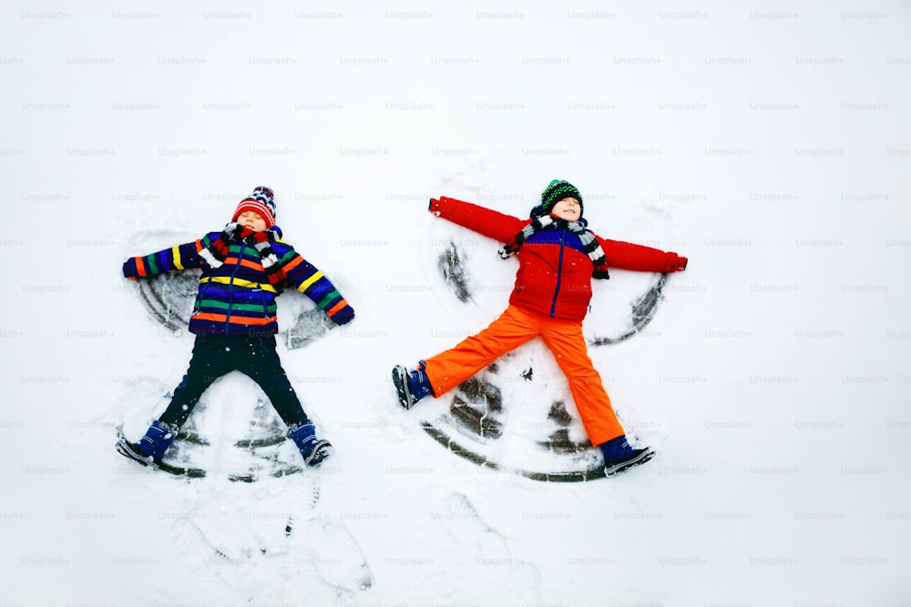 Two little siblings kid boys in colorful winter clothes making snow angel, laying down on snow. Active outdoors leisure with children in winter. Happy brothers with warm hat, gloves, winter fashion