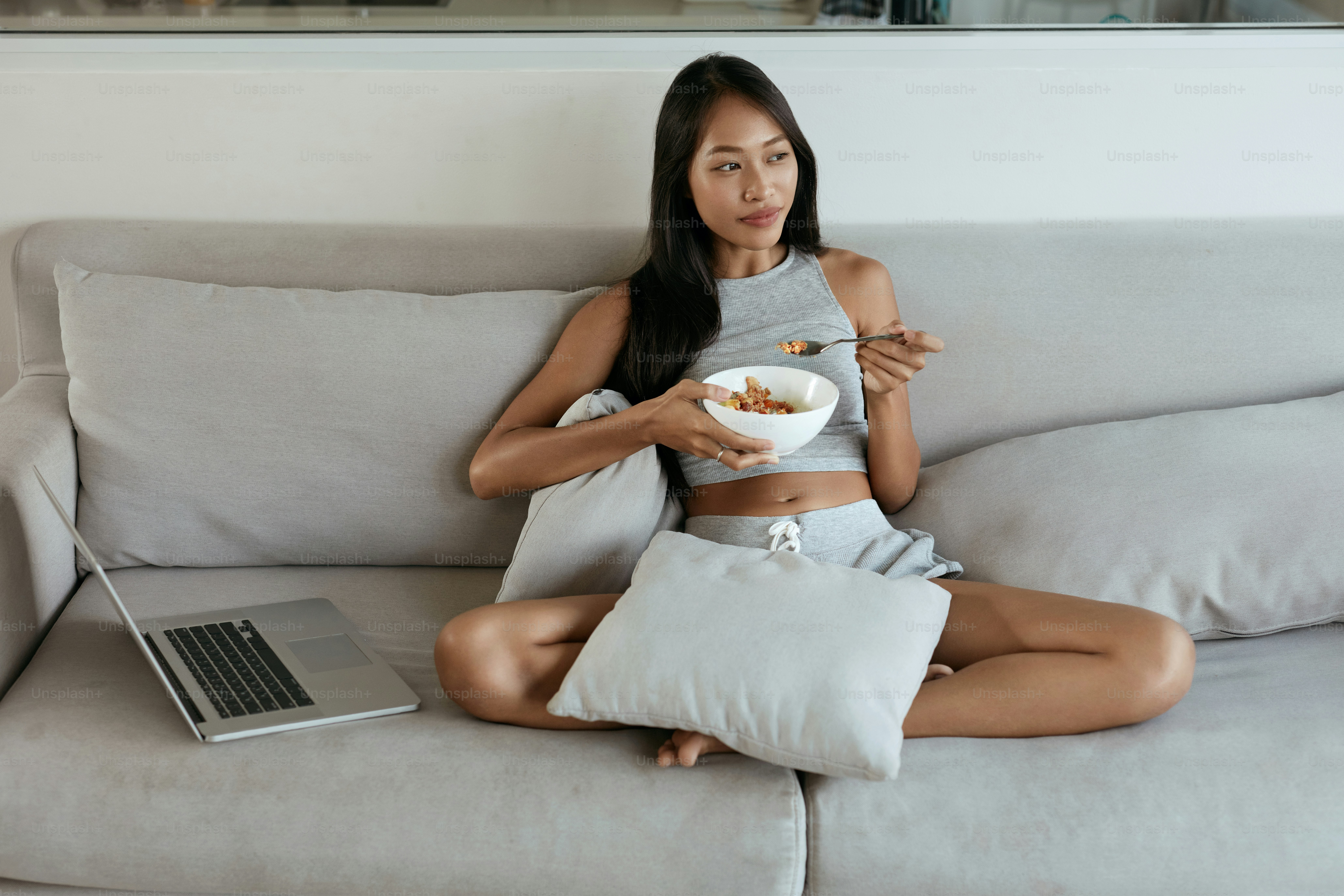 Mujer desayunando mientras usa la computadora en casa por la mañana. Hermosa chica asiática sonriente comiendo comida relajándose en el sofá con computadora portátil en la sala de estar luminosa