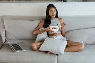 Woman having breakfast while using computer at home in morning. Beautiful smiling asian girl eating food relaxing on couch with laptop in light living room