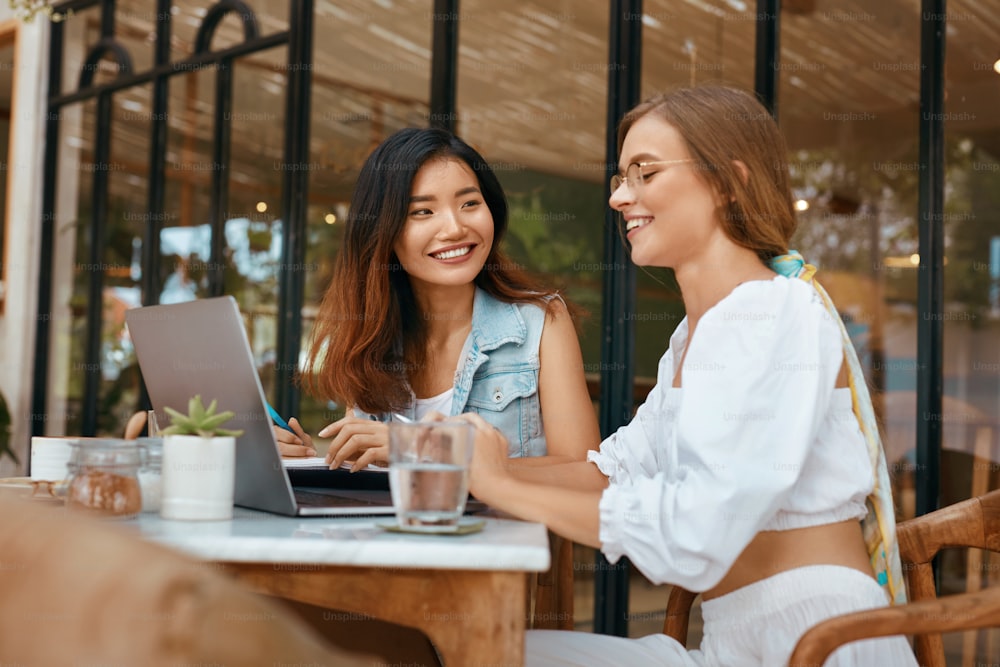 Women Working On Laptop At Cafe. Beautiful Ethnic Girls In Casual Clothes Having Discussion About Business On Terrace. Digital Nomad Lifestyle For Comfortable Work At Coffee Shop.