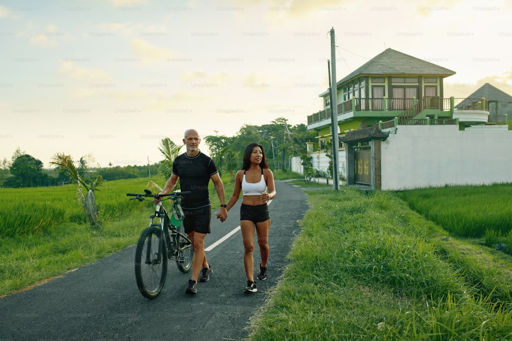 道路上の自転車を歩くスポーツなカップル。アジア人女性と白人男性が自転車で手をつないで歩き、緑の熱帯の風景を背景に話す。