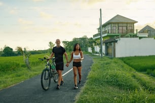 Coppia sportiva a piedi in bici su strada. Donna asiatica e uomo caucasico che cammina per mano con le biciclette e parla contro il paesaggio tropicale verde.