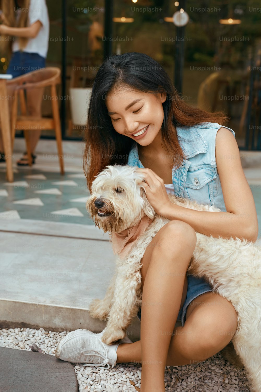 Asiatische Frau und Hund auf der Terrasse. Schönes Model in Jeans-Outfit spielt mit Welpe auf der Terrasse im hundefreundlichen Café. Glückliche Frau in modischer Kleidung umarmt Haustier und genießt Sommerurlaub.