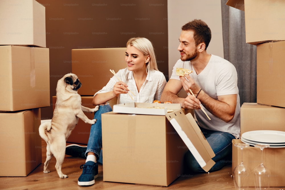 Spostarsi. Uomo felice e bella donna che mangiano vicino a scatole di cartone e cane in una nuova casa. Alta risoluzione.