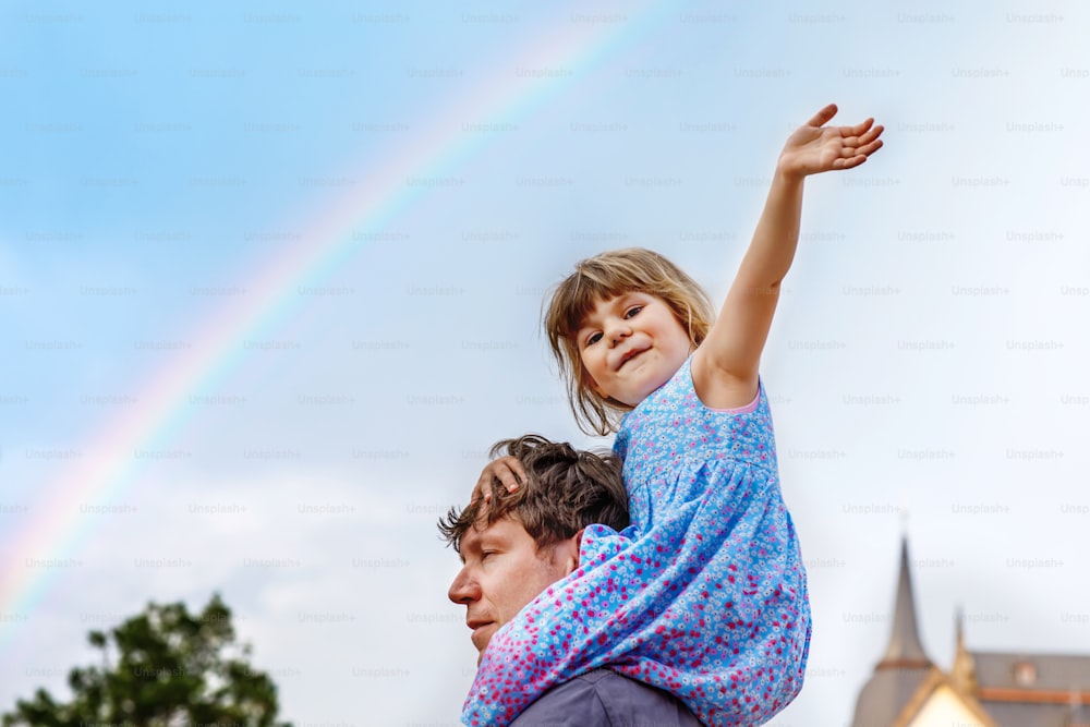 Little preschool girl sitting on shoulder of father. Happy toddler child and man observing rainbow on sky after summer rain. Happy family, bonding, love. Summertime. Dad and daughter. Father's day