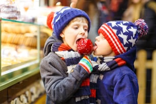 두 명의 어린 소년, 귀여운 형제 자매가 진저브레드와 견과류를 들고 달콤한 스탠드 근처에서 설탕 사과를 먹고 있습니다. 독일의 크리스마스 시장에서 행복한 아이들. 크리스마스의 전통적인 여가.