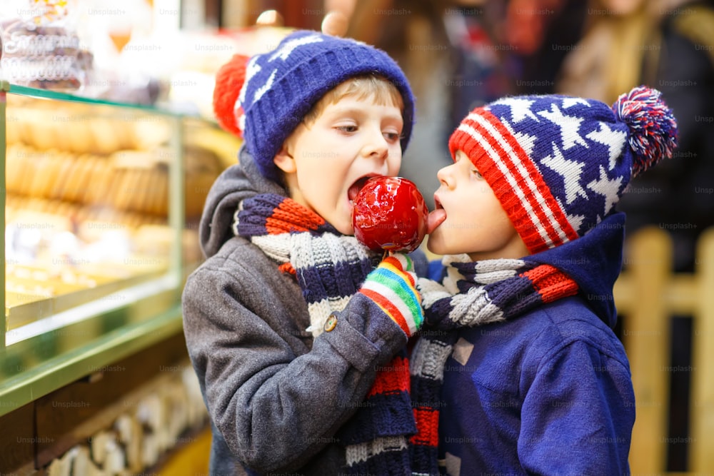 Dois meninos pequenos, irmãos bonitos comendo maçã de açúcar perto de carrinho doce com pão de gengibre e nozes. Crianças felizes no mercado de Natal na Alemanha. Lazer tradicional no Natal.
