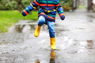 Gros plan d’un enfant portant des bottes de pluie jaunes et marchant pendant le grésil, la pluie et la neige par temps froid. Enfant dans des vêtements décontractés colorés sautant dans une flaque d’eau. S’amuser à l’extérieur.