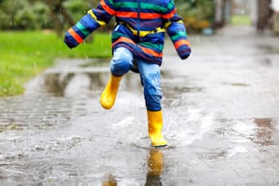 Close-up of kid wearing yellow rain boots and walking during sleet, rain and snow on cold day. Child in colorful fashion casual clothes jumping in a puddle. Having fun outdoors.