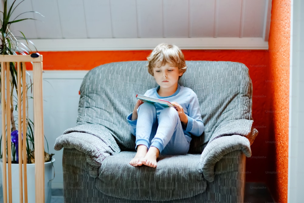 Lindo niño rubio en preescolar en pijama leyendo libro para niños en la habitación doméstica. Niño emocionado leyendo en voz alta, sentado en un viejo sillón grande. Escolar, familia, educación.