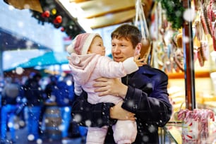 ジンジャーブレッドとナッツを使った甘いスタンドの近くに赤ん坊の娘を抱えた中年の父親。ドイツのクリスマスマーケットで幸せな家族。レープクーヘンというクッキーを食べるかわいい女の子。お祝いのクリスマス休暇