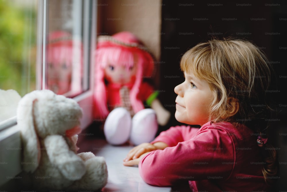 Cute toddler girl sitting by window and looking out on rainy day. Dreaming child with doll and soft toy feeling happy. Self isolation concept during corona virus pandemic time. Lonely kid