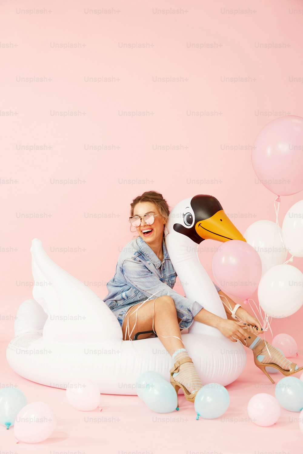 Mujer de moda en ropa de verano divirtiéndose con globos. Chica sonriente feliz en ropa de moda y gafas de sol elegantes con globos de colores sentados en flotador de cisne inflable blanco en el fondo rosa