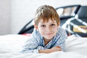 Adorable happy little kid boy after sleeping in his white bed in colorful nightwear. School child celebrating pajama party and looking at the camera. Funny happy child playing and smiling.