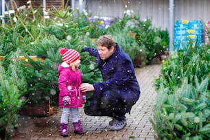 Little toddler girl and father holding Christmas tree on a market. Happy family, cute baby child daughter and middle aged man in winter fashion clothes choosing and buying Xmas tree in outdoor shop