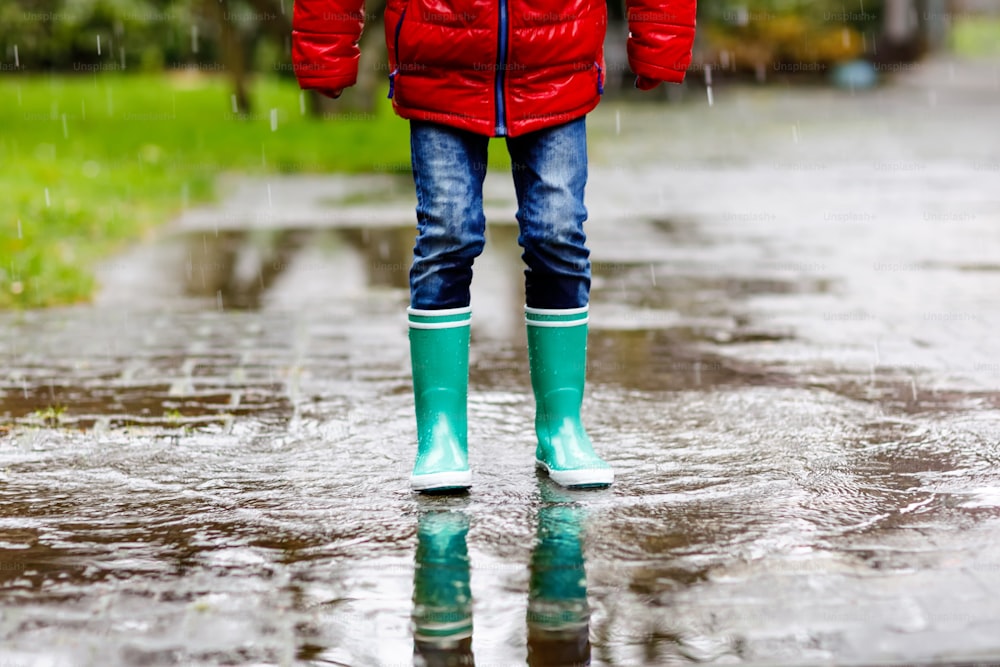 Close-up of kid wearing yellow rain boots and walking during sleet, rain and snow on cold day. Child in colorful fashion casual clothes jumping in a puddle. Having fun outdoors.