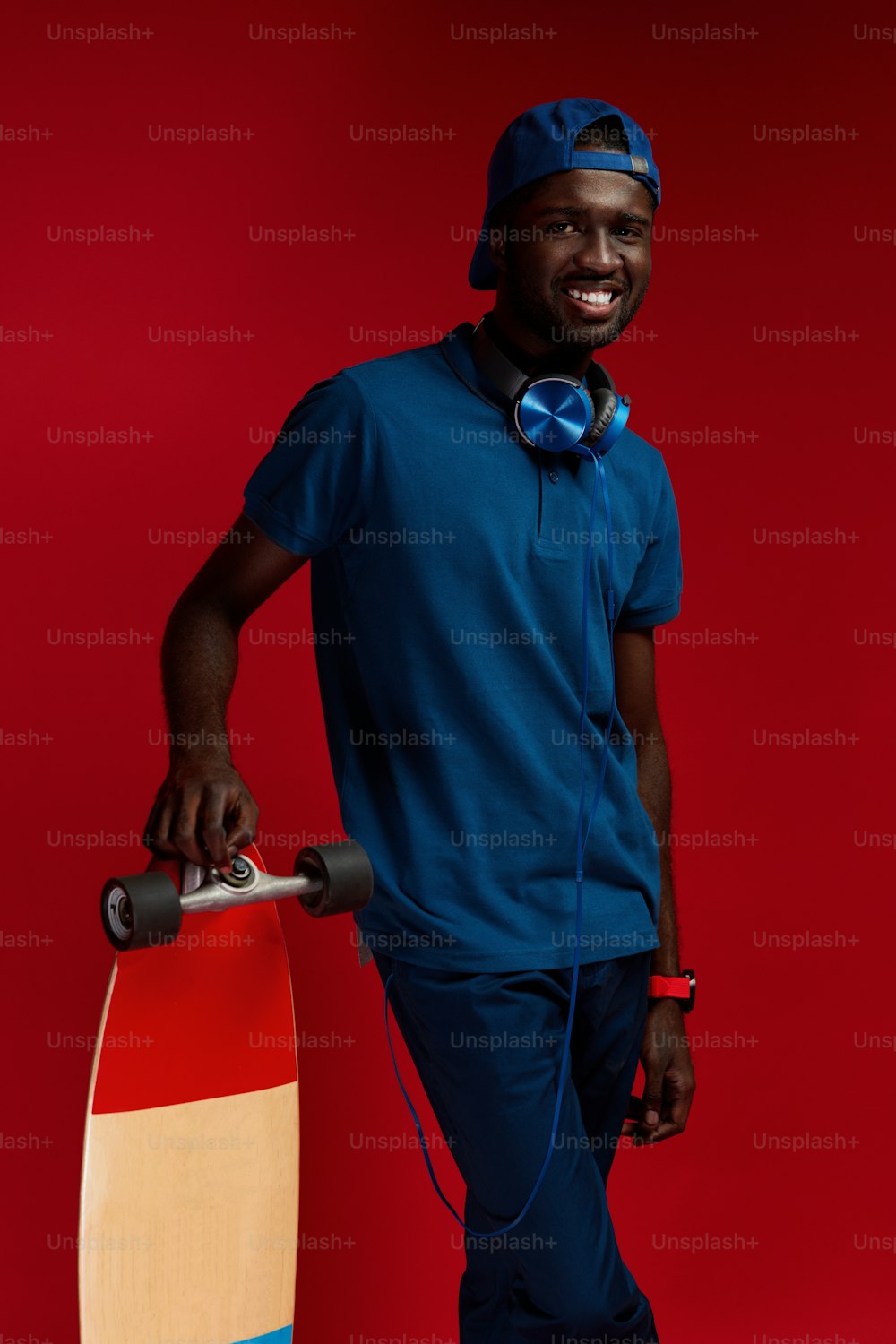 Fashion. Smiling african man in stylish outfit with skateboard at studio on red background. Happy black male model in blue cap, polo shirt with headphones on neck and skate board