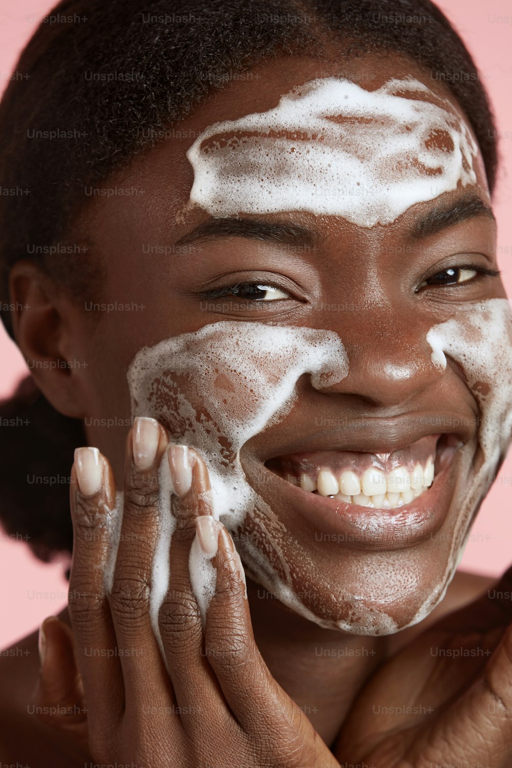 Ritratto ravvicinato della bella ragazza nera lavarsi il viso con schiuma detergente per il viso. Giovane donna sorridente che guarda la macchina fotografica. Concetto di cura della pelle del viso. Isolato su sfondo rosa. Riprese in studio