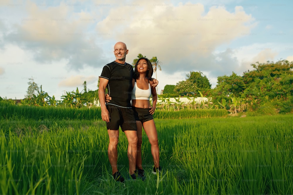 Coppia matura in piedi sul ritratto del campo di riso. Uomo caucasico e donna asiatica che riposano dopo l'allenamento al mattino contro il paesaggio tropicale.