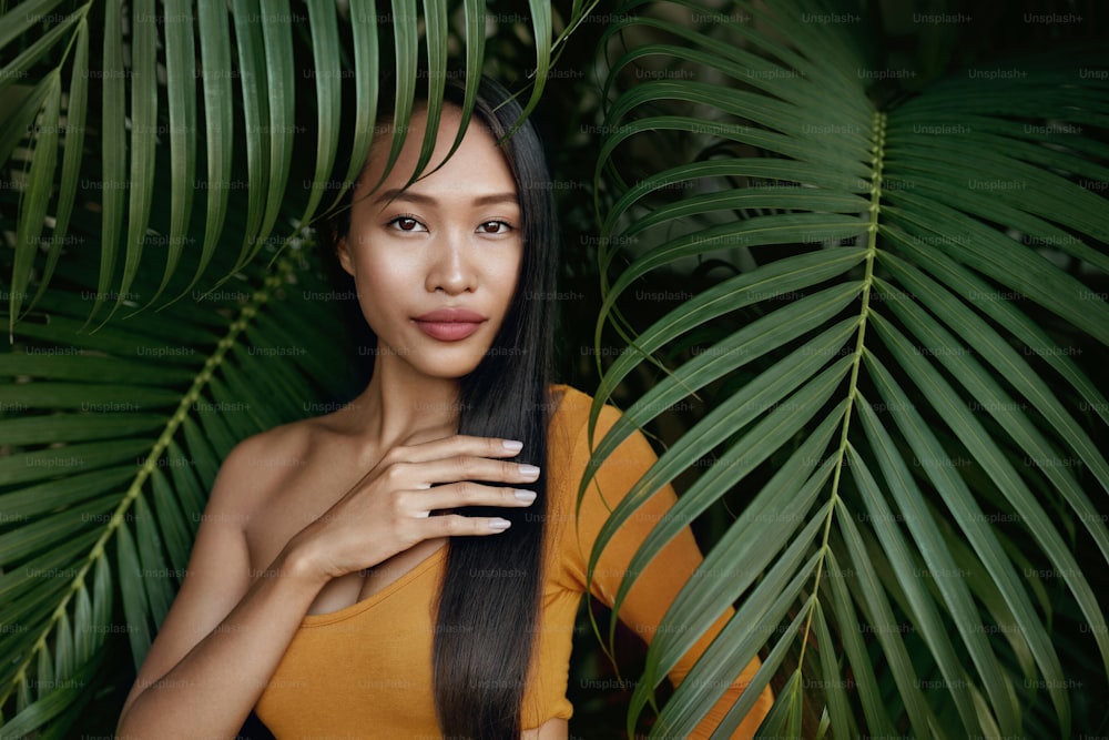 Hair beauty. Woman model with long straight hair in palm leaves. Beautiful asian girl with exotic face and natural makeup at tropical nature
