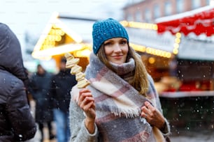 Beautiful young woman eating white chocolate covered fruits on skewer on traditional German Christmas market. Happy girl on traditional family market in Germany, Munich during snowy day