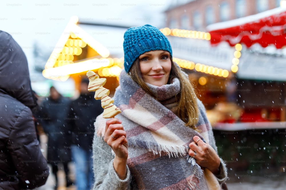 Schöne junge Frau, die auf dem traditionellen deutschen Weihnachtsmarkt mit weißer Schokolade überzogene Früchte am Spieß isst. Glückliches Mädchen auf traditionellem Familienmarkt in Deutschland, München an einem verschneiten Tag