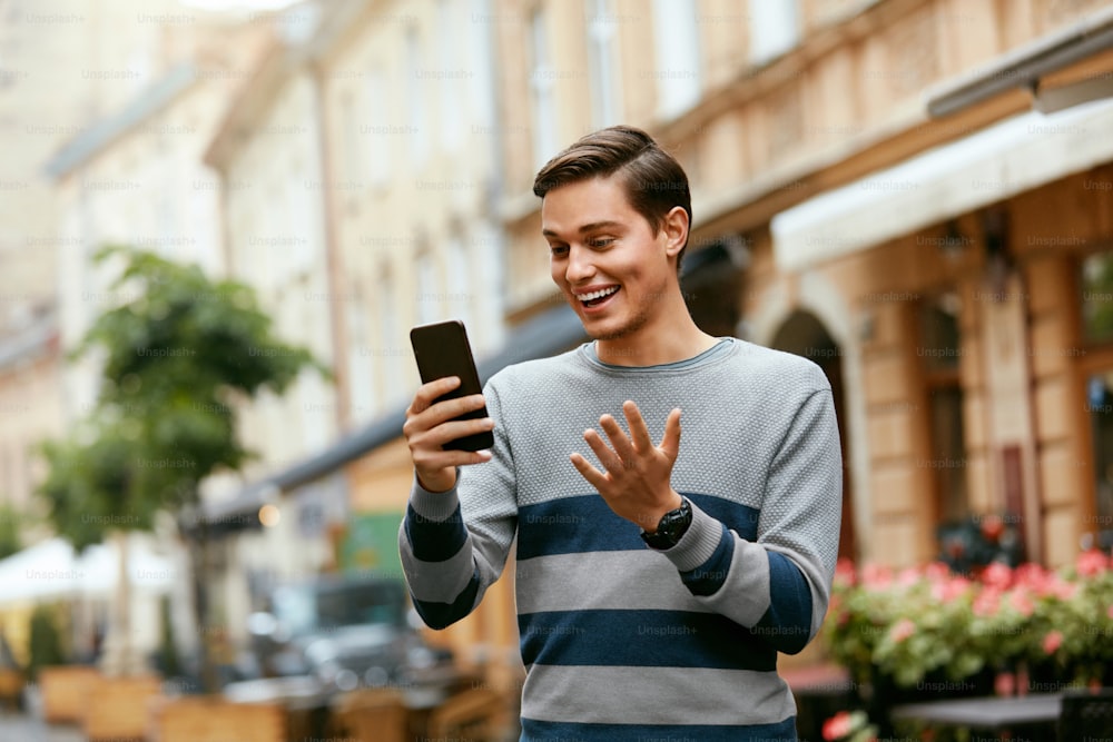 Man Video Calling On Phone On Street. Smiling Young Male Using Mobile Phone Outdoors. High Resolution