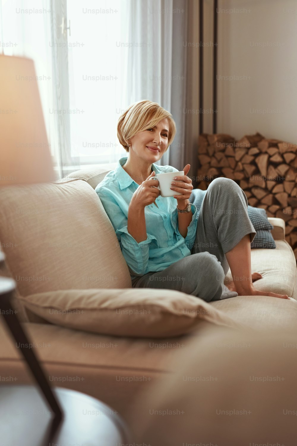 Mature Woman. Beautiful Healthy Middle Age Female Sitting On Sofa At Home. Model Drinks From Cup. Beauty And Comfort Concept.