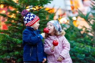 Dos hermanos sonrientes, niños comiendo manzana azucarada cristalizada en el mercado navideño alemán. Niños felices, gemelos en ropa de invierno con luces de fondo. Familia, tradición, concepto vacacional