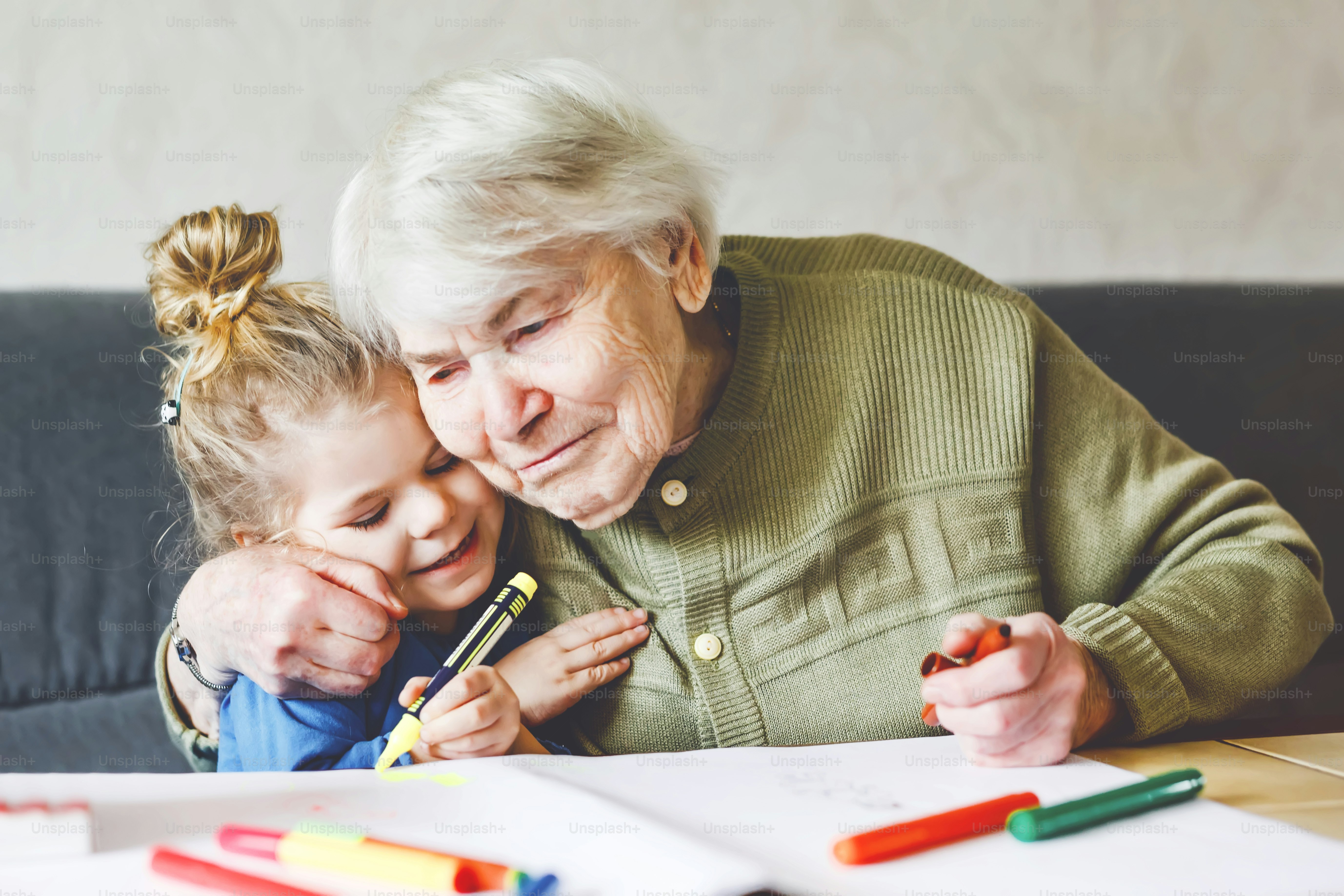 Beautiful toddler girl and grand grandmother drawing together pictures with felt pens at home. Cute child and senior woman having fun together. Happy family indoors.