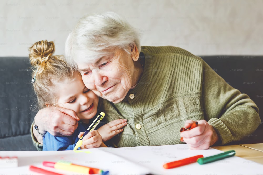 Beautiful toddler girl and grand grandmother drawing together pictures with felt pens at home. Cute child and senior woman having fun together. Happy family indoors.