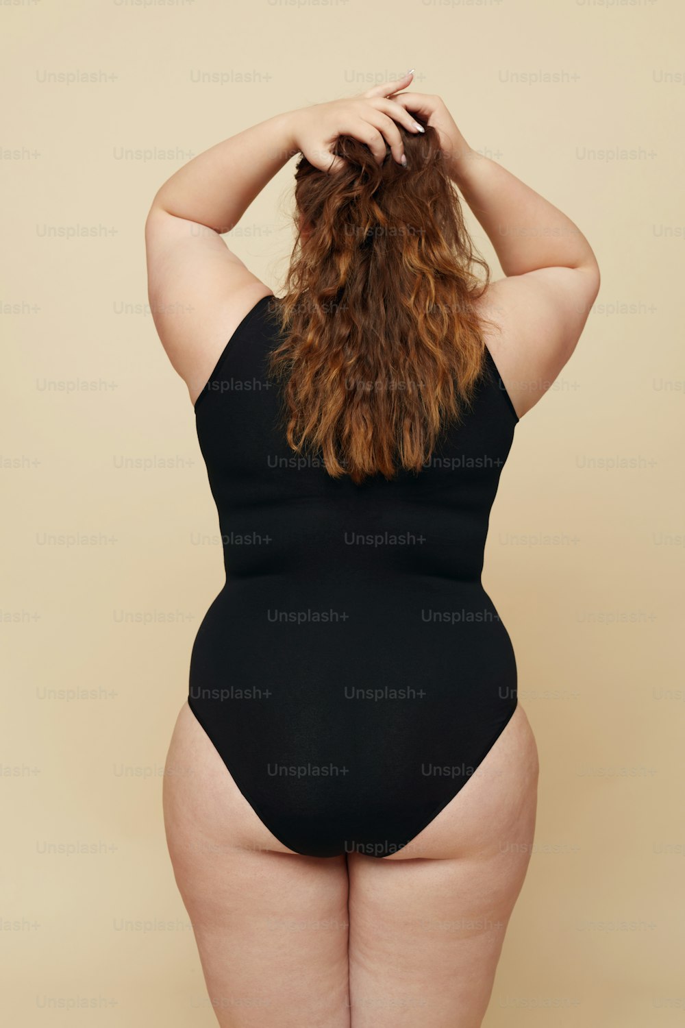 Plus Size Model. Woman From Back Portrait. Full-figured Brunette In Back Bodysuit Keeping Hands On Head And Touching Hair. Body Positive Concept On Beige Background.