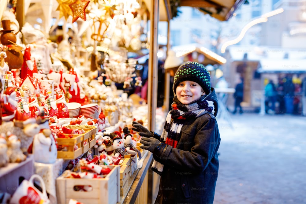 クリスマスマーケットで飾りを選ぶ小さなかわいい男の子。おもちゃや木の装飾的な装飾品を買う美しい子供。ドイツのクリスマスマーケット