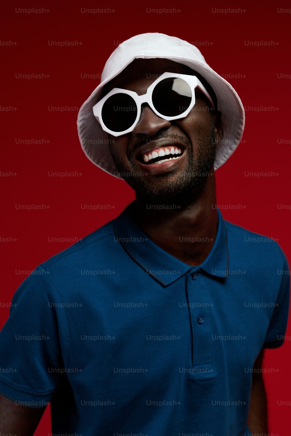 赤い背景に白い帽子とサングラスをかけた笑顔の黒人男性のファッションポートレート。スタジオでスタイリッシュな夏の衣装とアイウェアを着た幸せなアフリカ系アメリカ人男性モデル