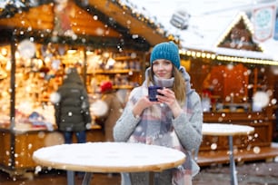 Schöne junge Frau, die heißen Punsch und Glühwein auf dem deutschen Weihnachtsmarkt trinkt. Glückliches Mädchen in Winterkleidung mit Lichtern auf dem Hintergrund am Winterschneetag in München, Deutschland