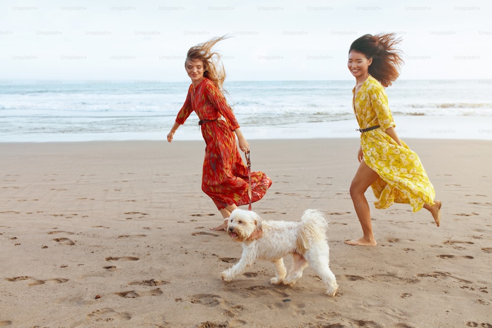 Été. Filles avec chien sur la plage. Femmes de mode en robes bohèmes courant pieds nus avec animal de compagnie sur un complexe acceptant les chiens. Des modèles heureux en tenue tendance en vacances à l’océan tropical.