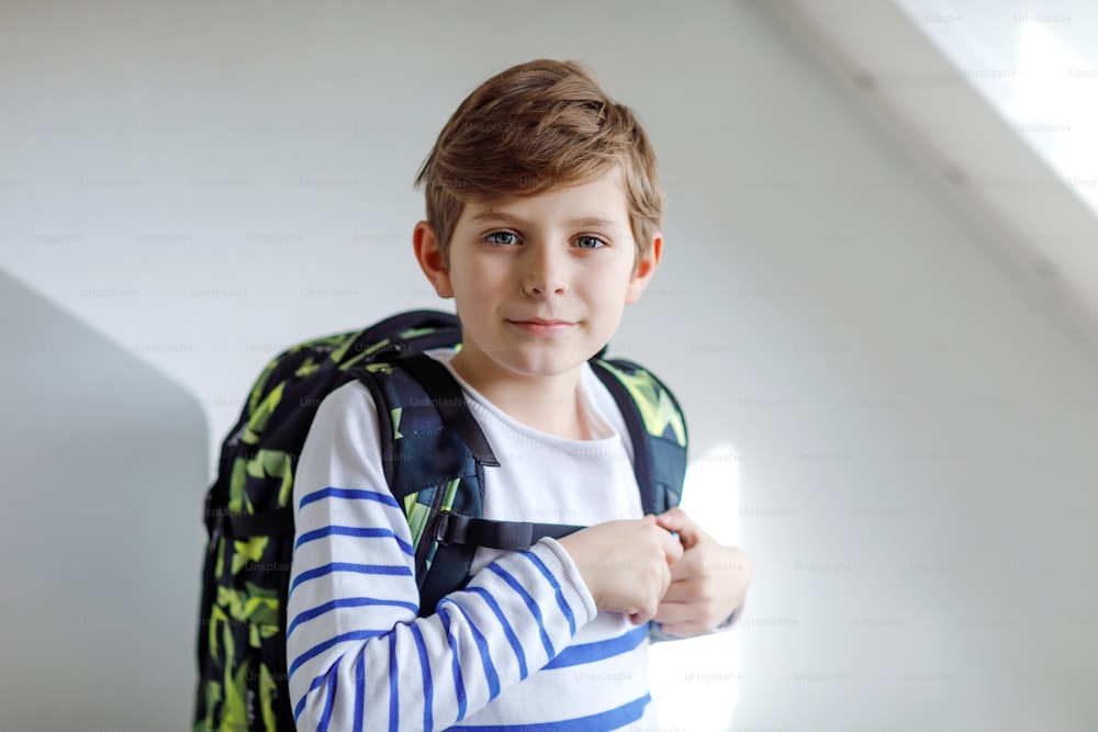 Beautiful kid boy with schoolbag rucksack standing in room in early morning. Happy healthy child on the way to lessons and school. Education concept.
