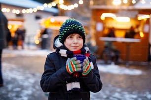 Garotinho bonito menino bebendo soco de crianças quentes ou chocolate no mercado de Natal alemão. Criança feliz no mercado tradicional da família na Alemanha, menino rindo em roupas coloridas de inverno.