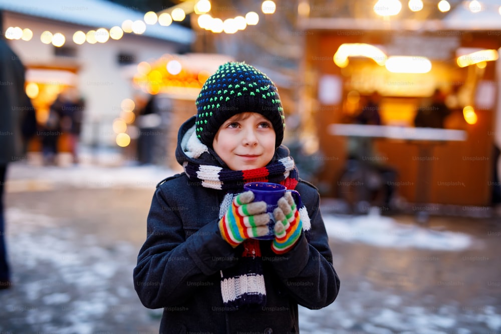 Little cute kid boy drinking hot children punch or chocolate on German Christmas market. Happy child on traditional family market in Germany, Laughing boy in colorful winter clothes.