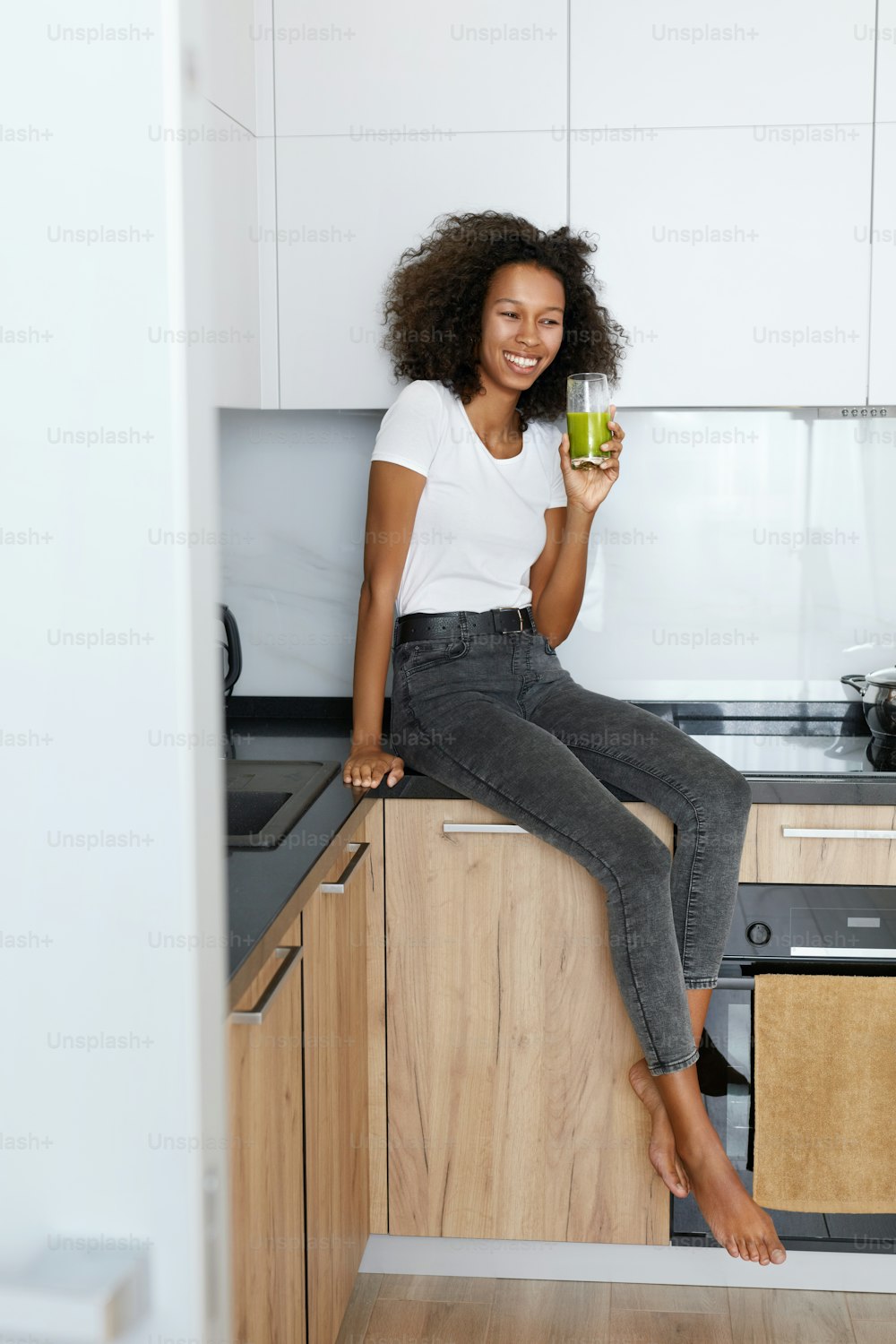 Woman drinking green detox juice, smoothie drink in kitchen. Happy smiling african girl with glass of healthy fresh raw vegetable smoothie at home. Diet nutrition concept