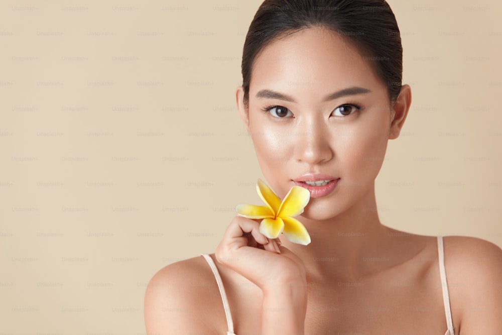 Beauté. Portrait de femme et de fleur. Le modèle asiatique tient le plumeria tropical près du visage et regarde la caméra sur fond beige. Belle fille ethnique avec une peau faciale parfaite, éclatante et hydratée.