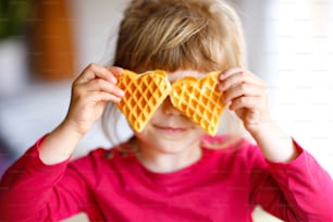 Retrato da menina pré-escolar feliz segurando waffle de coração fresco assado. Criança criança faminta sorridente com bolacha de biscoito doce. Waffles belgas de açúcar doce