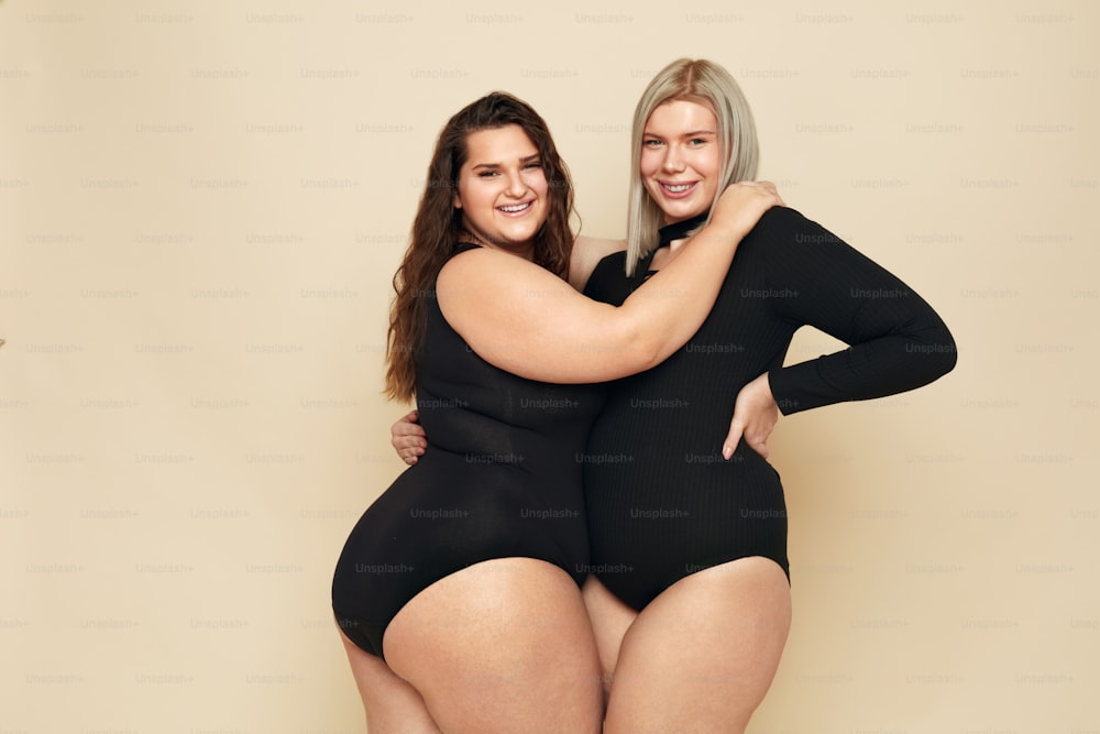 Plus Size Models. Full-figured Women Portrait. Brunette And Blonde In Black Bodysuits Posing On Beige Background. Smiling Female Looking At Camera. Body Positive As Lifestyle.