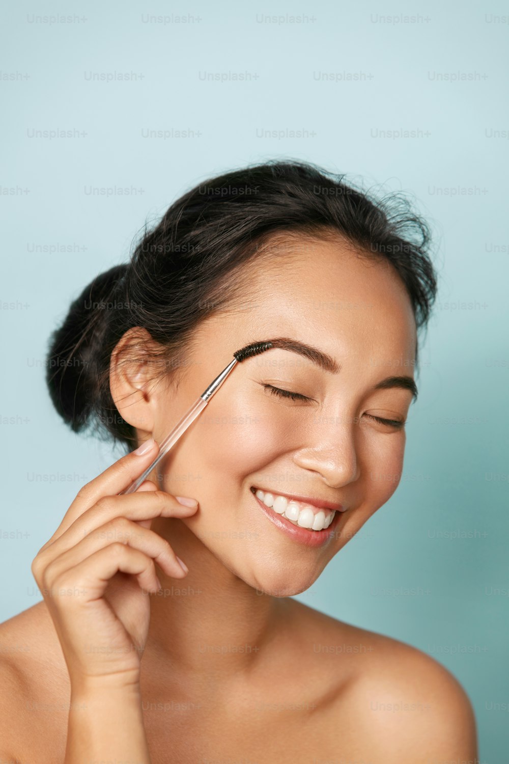Belleza. Mujer sonriente cepillando las cejas con primer plano del pincel de maquillaje. Retrato de una hermosa modelo asiática feliz maquillándose, dando forma a las cejas con un pincel cosmético en el estudio.
