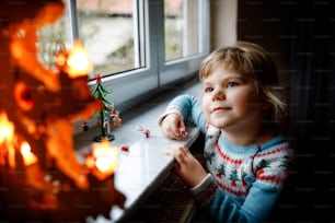 Litte toddler girl sitting by window and decorating small glass Christmas tree with tiny xmas toys. Happy healthy child celebrate family traditional holiday. Adorable baby